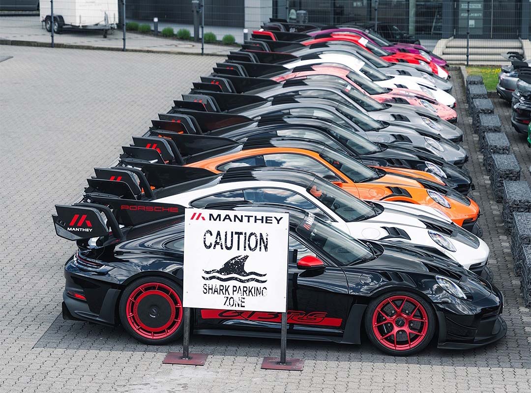 A row of parked Porsche sports cars with large rear wings, including a GT3 RS MR, sits behind a sign reading "Manthey Caution Shark Parking Zone," showcasing Manthey Racing’s race-ready machines.
