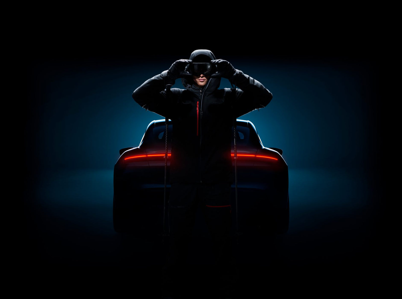 A person in dark clothing and a helmet stands in front of a Porsche sports car with illuminated rear lights, set against a dark background.