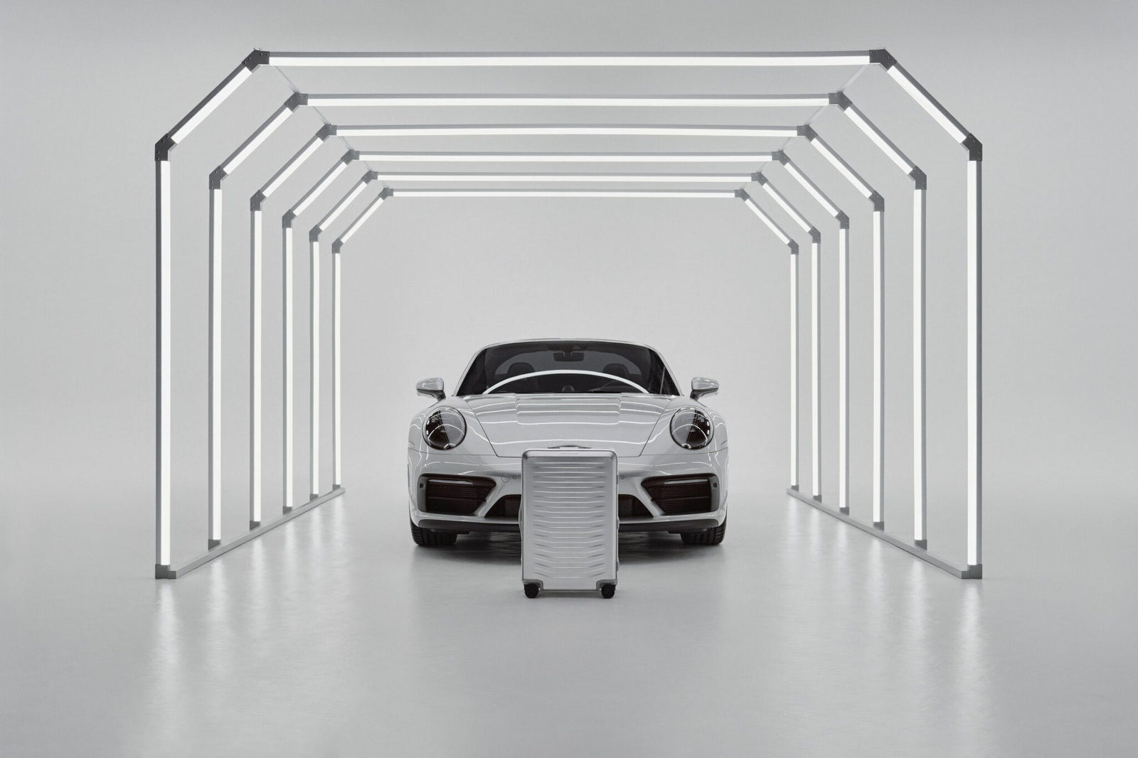 A white sports car is parked inside a minimalistic light tunnel, with a Porsche Design Roadster Aluminum Luggage suitcase in matching white positioned in front—an exquisite scene that inspires you to buy your next travel essential.
