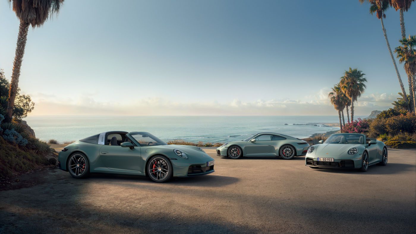 Three silver Porsche 911 variants are parked on a coastal road lined with palm trees, overlooking the ocean beneath a clear sky.