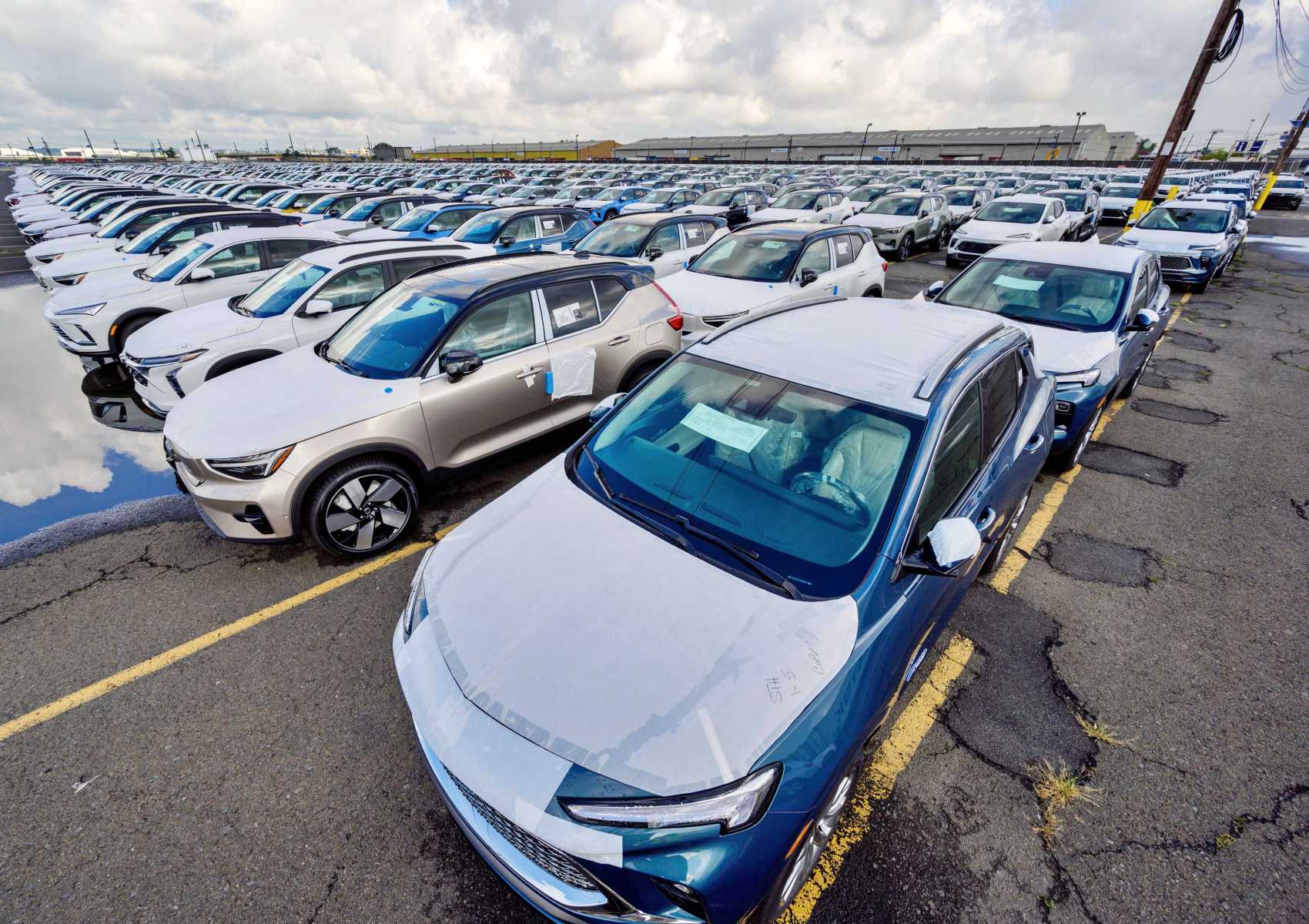 Rows of new cars are parked in a large outdoor lot on an overcast day, with puddles reflecting the vehicles—poised for export as the Trump von der Leyen landmark deal aims to boost U.S. auto exports to Europe by slashing tariffs.