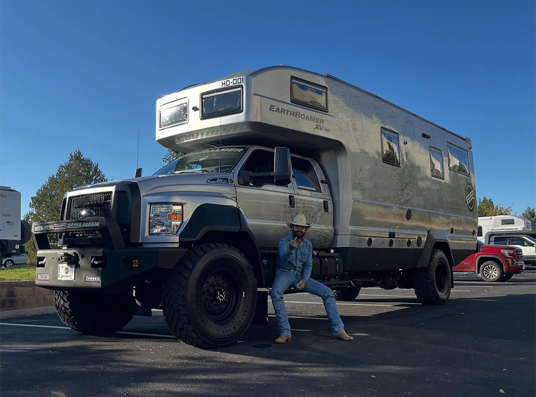 A person in a hat and jeans sits on the front bumper of a large off-road EarthRoamer HD RV parked in a lot on a clear day.