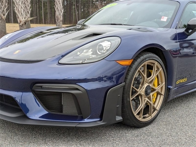 A close-up of the front left side of a Gentian Blue Porsche 718 Cayman GT4 RS sports car, featuring the headlight, wheel, and part of the hood with highlights from the Weissach Package.