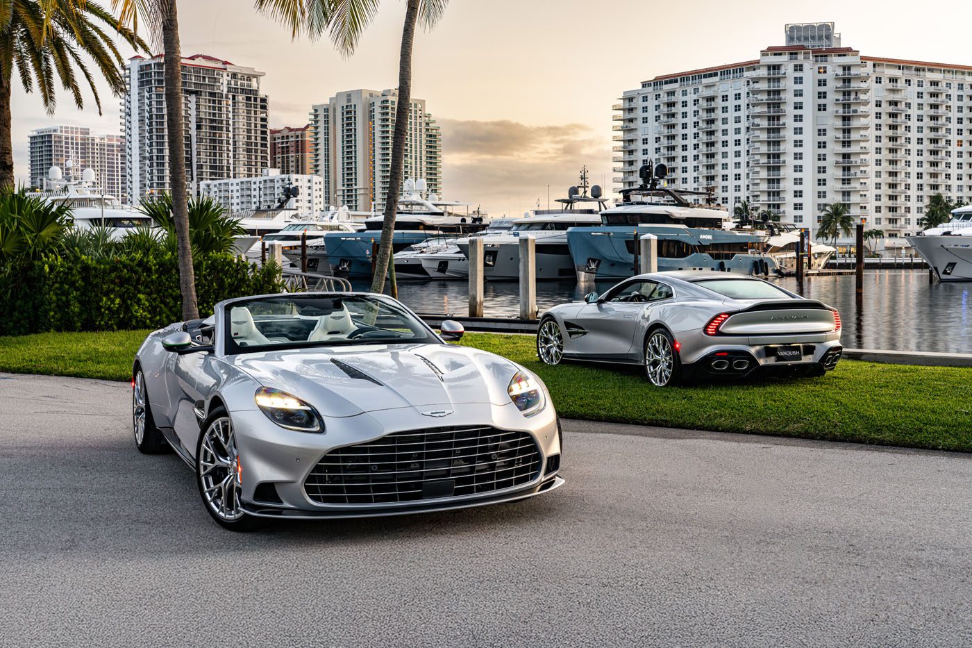 Two silver convertible sports cars are parked near a marina with yachts and high-rise buildings in the background.