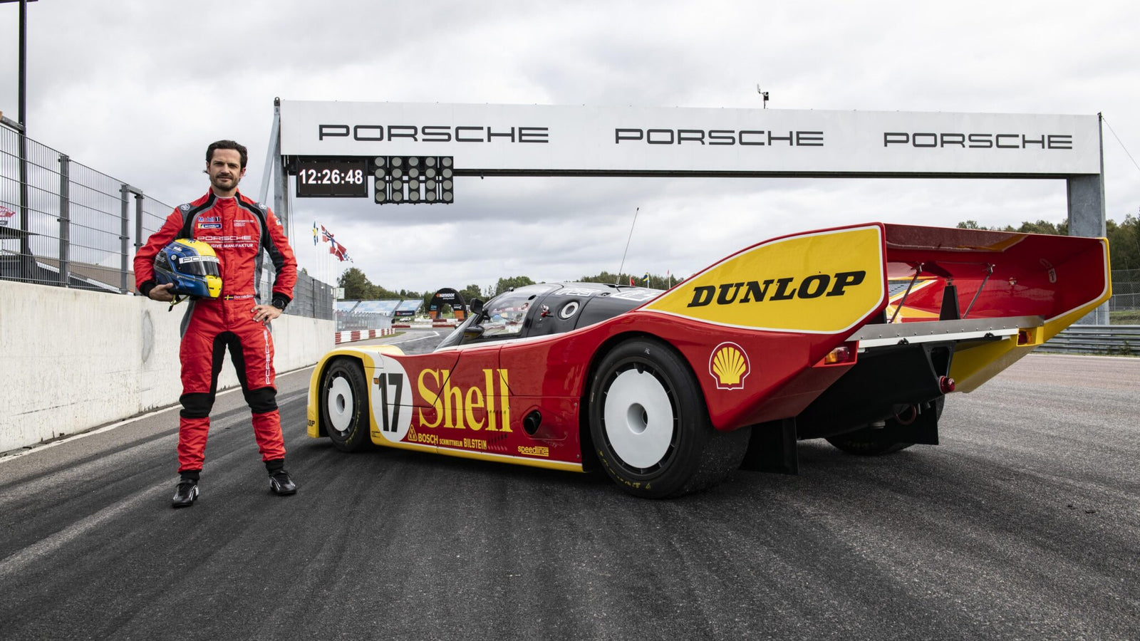 A person in a red racing suit stands next to a vintage Porsche 962 C with Shell and Dunlop branding on the track at Mantrop Park, beneath a Porsche sign.