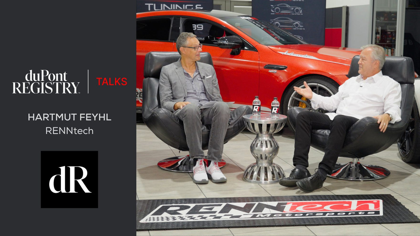 Two men in conversation sit in chairs with a table between them, a red AMG car in the background, and duPont REGISTRY and RENNtech branding displayed.