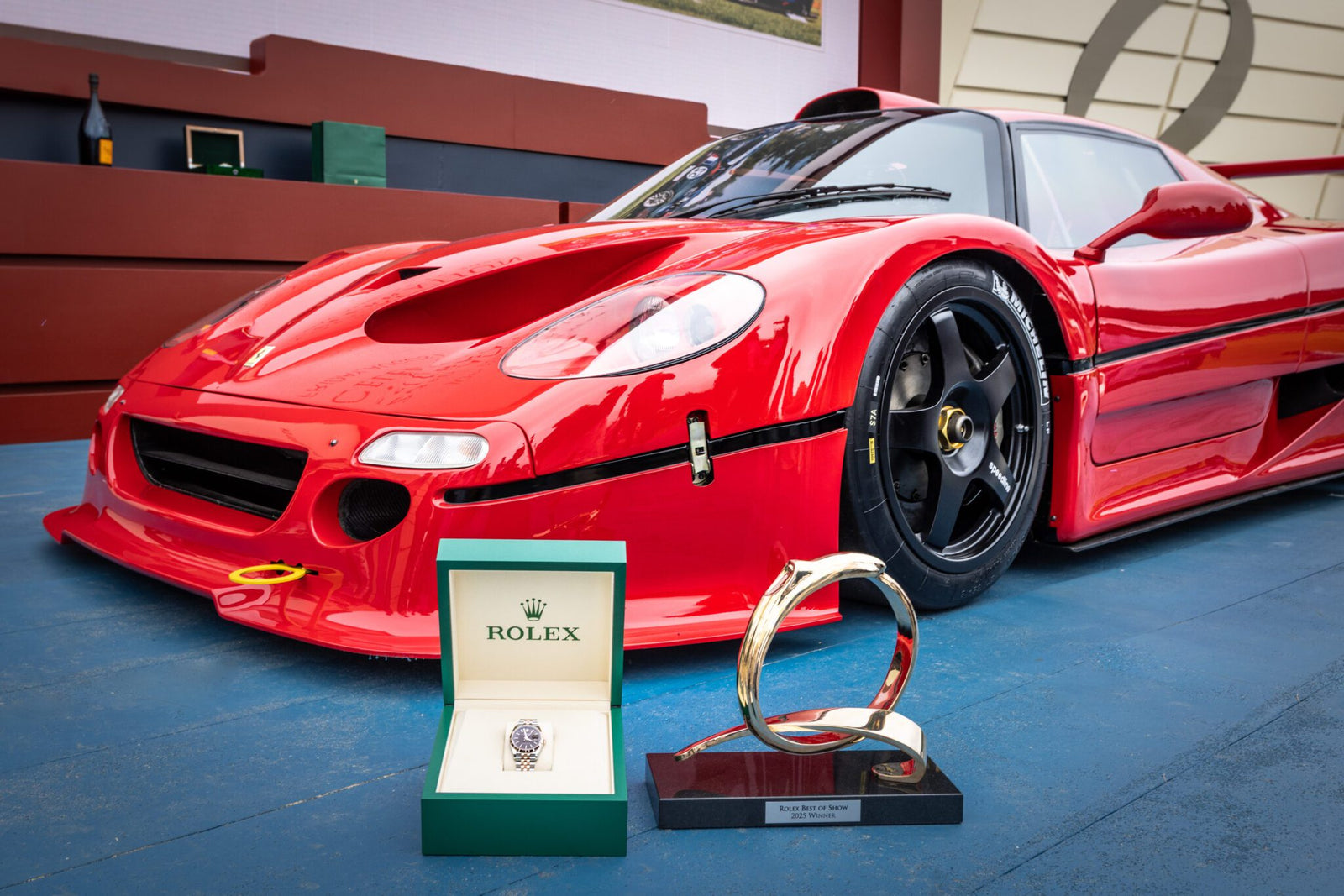 A red sports car is parked beside a luxury watch in an open box and a circular trophy on a stand, capturing the elegance of Monterey Car Week.