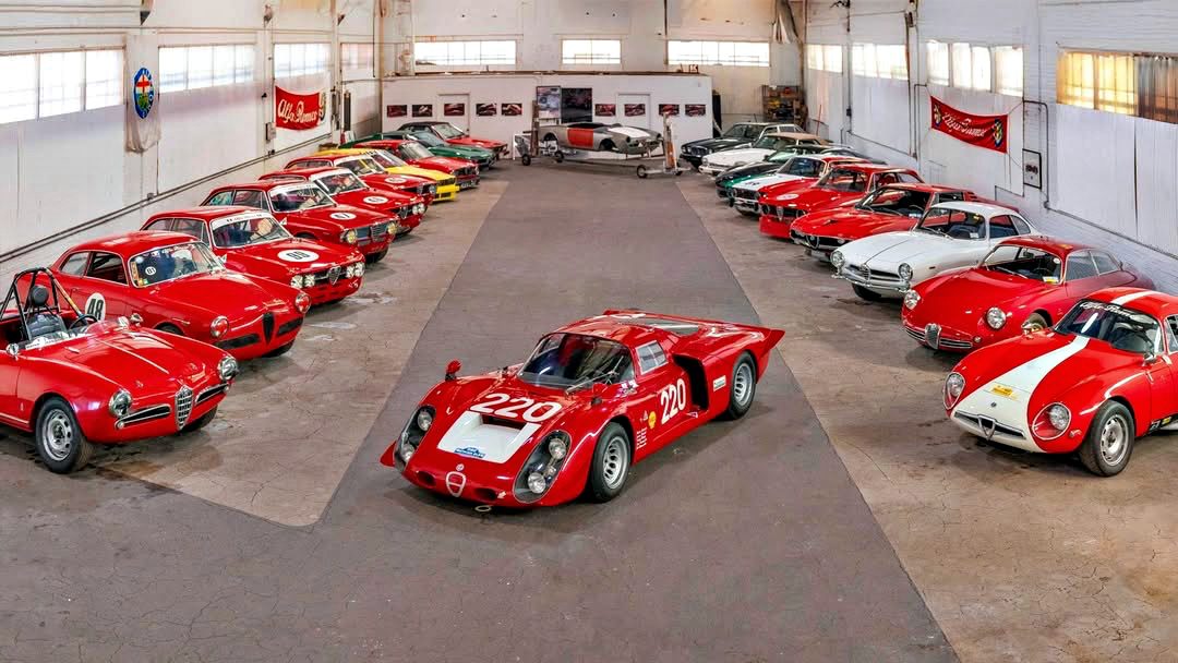 A collection of classic sports cars, mostly red, arranged in a large garage with a race car featuring number 220 in the center, showcased as part of the RM Sotheby’s Quadrifoglio Collection Monterey 2025.