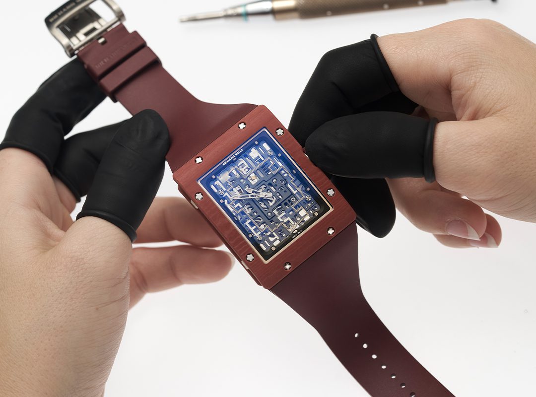 Person wearing black gloves is holding a red RM 16-02 from the Richard Mille Extraflat Collection, featuring a transparent dial that reveals its intricate internal components. A screwdriver is blurred in the background.