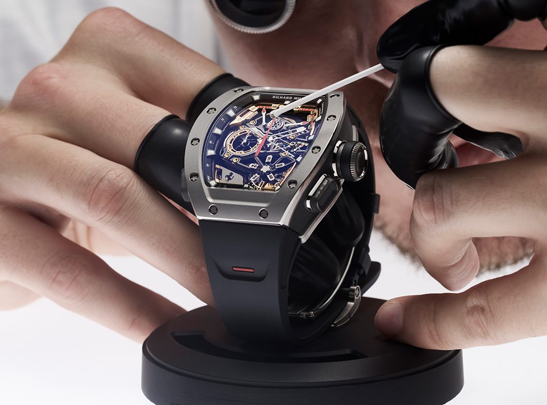 A person wearing black gloves is meticulously inspecting a Richard Mille luxury watch with a blue face and visible gears, carefully using a tool while it's mounted on a stand.
