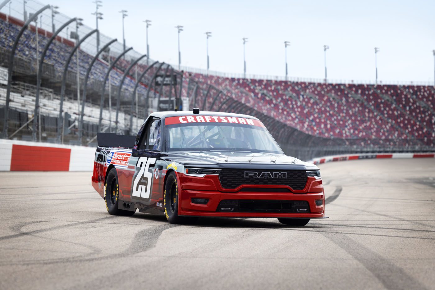 A black and red Ram race truck with the number 25 on the side, powered by HEMI V8s, drives on a racetrack with empty grandstands in the background.