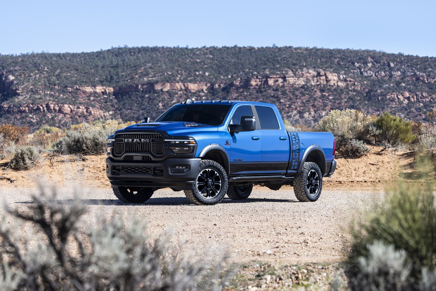A blue 2027 Ram Power Wagon pickup truck is parked on a dirt road in a desert landscape with shrubs and rocky hills in the background, ready to showcase its impressive torque.