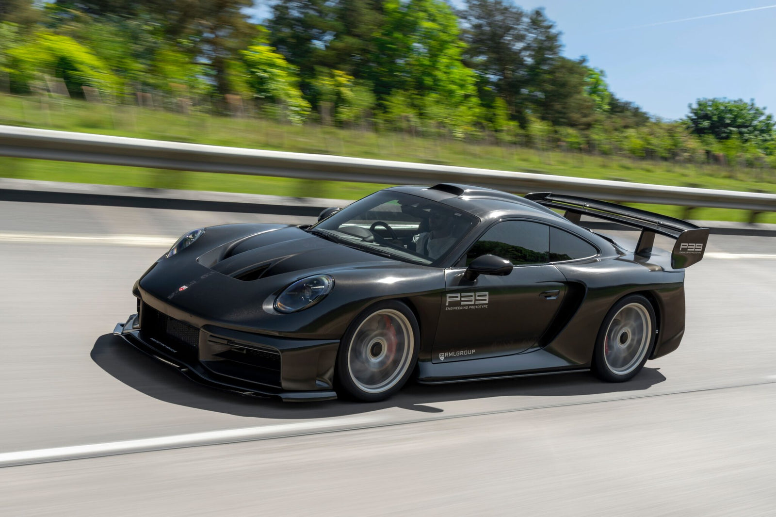 A matte black Porsche sports car with a large rear wing drives on a highway, surrounded by greenery and trees—a scene reminiscent of an RML Group P39 Hypercar or a Graham Rahal Performance masterpiece from a premier Hypercar Dealer.