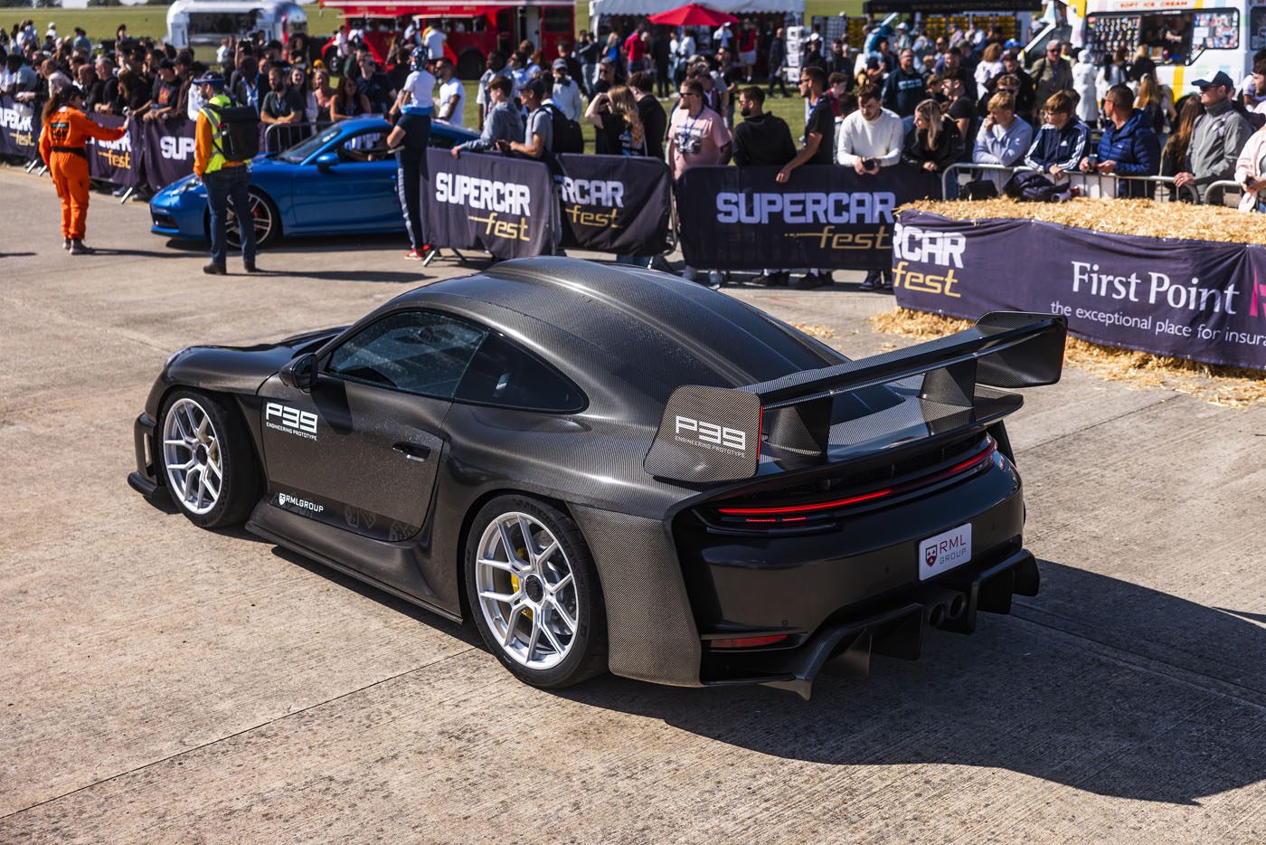 A black sports car with a large rear wing is displayed at an outdoor car event, surrounded by spectators and other parked cars.