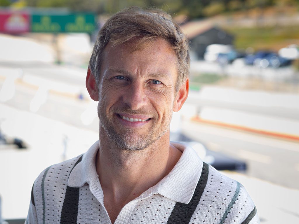 A man with light brown hair and a light beard smiles while standing outdoors on a sunny day, wearing a white and green collared shirt. The blurred road hints at the excitement of the Rolex Monterey Motorsports Reunion with Grand Marshal Jenson Button.