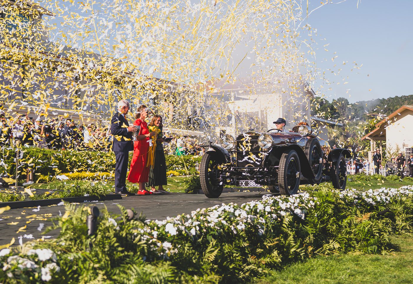A vintage Hispano-Suiza drives on stage surrounded by yellow confetti, as people applaud nearby at the Pebble Beach Concours d’Elegance after winning Best of Show at this prestigious outdoor event.