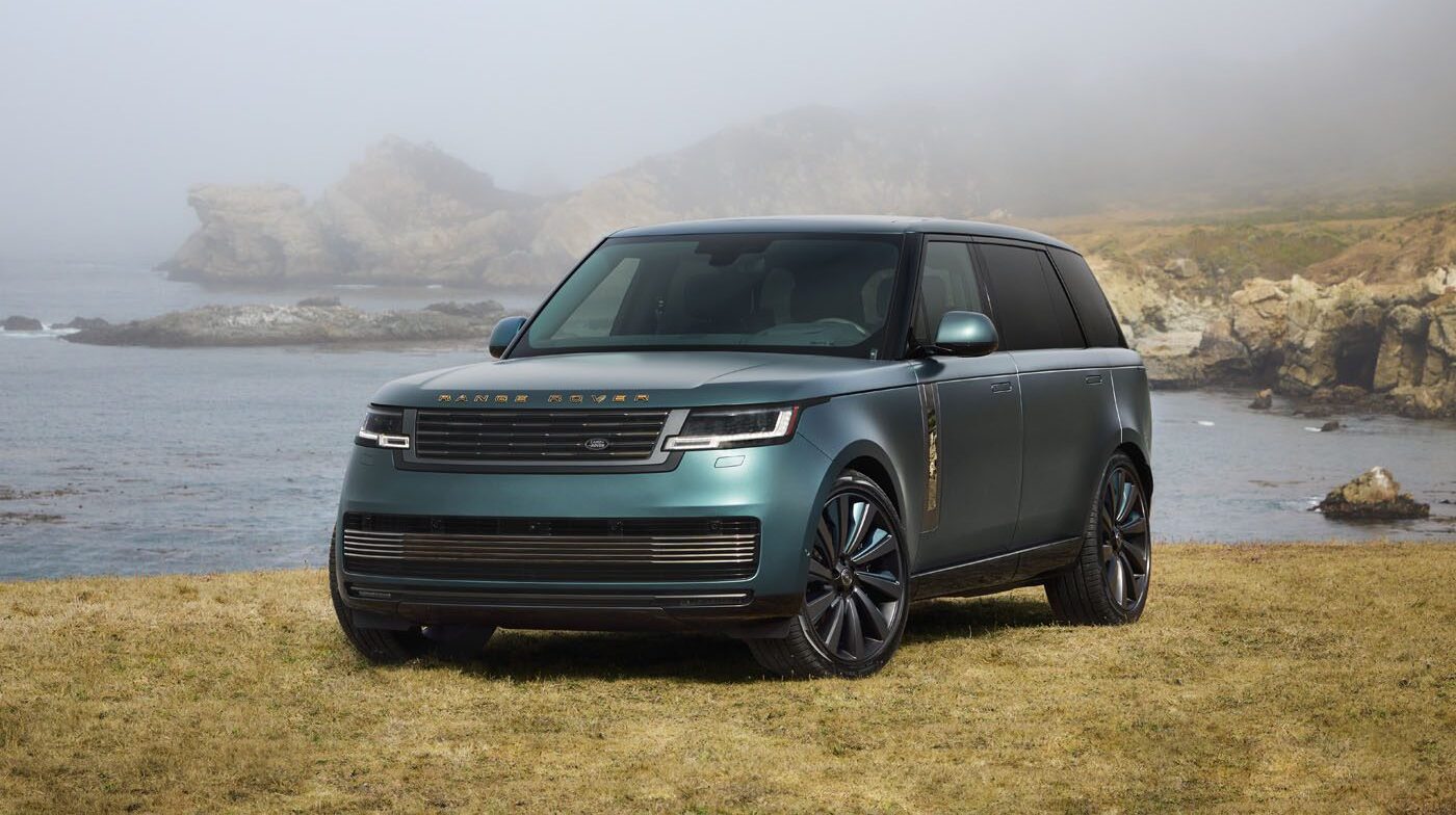 A green Range Rover SV Carmel Edition is parked on grass near a rocky shoreline, with fog and hills in the background—a true highlight among Exclusive Rovers.