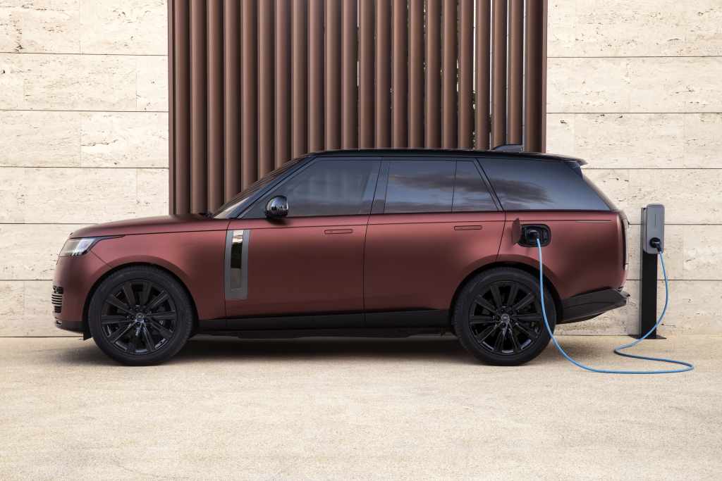 A maroon electric SUV from JLR is parked and plugged into a charging station against a beige wall, highlighting the shift towards hybrid sales in anticipation of FY25.