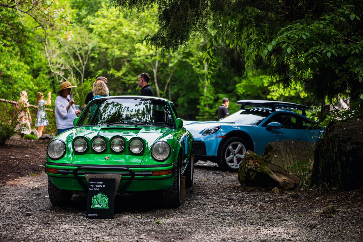 A green vintage Porsche sits elegantly outdoors beside a sleek blue sports car, drawing admirers into animated plática amid the shade of towering trees at this vibrant event.