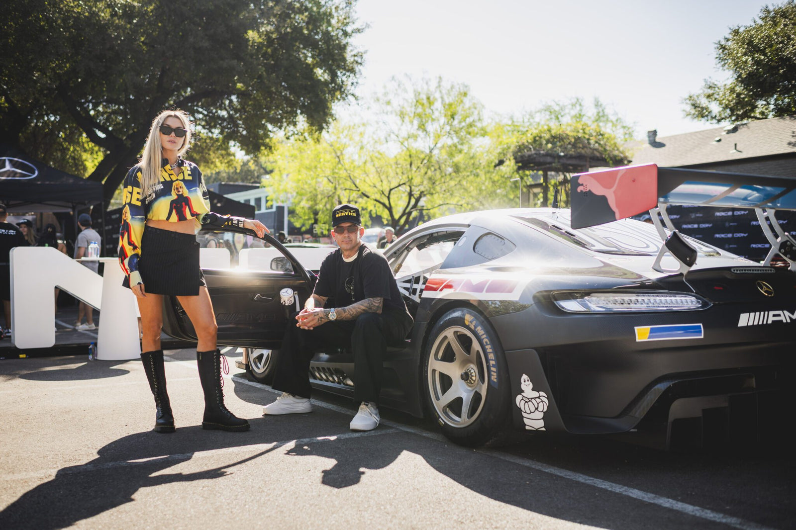 A woman and a man are posing beside a sleek Mercedes-Benz sports car, set against the vibrant backdrop of trees and event tents at the United States Grand Prix in Austin.