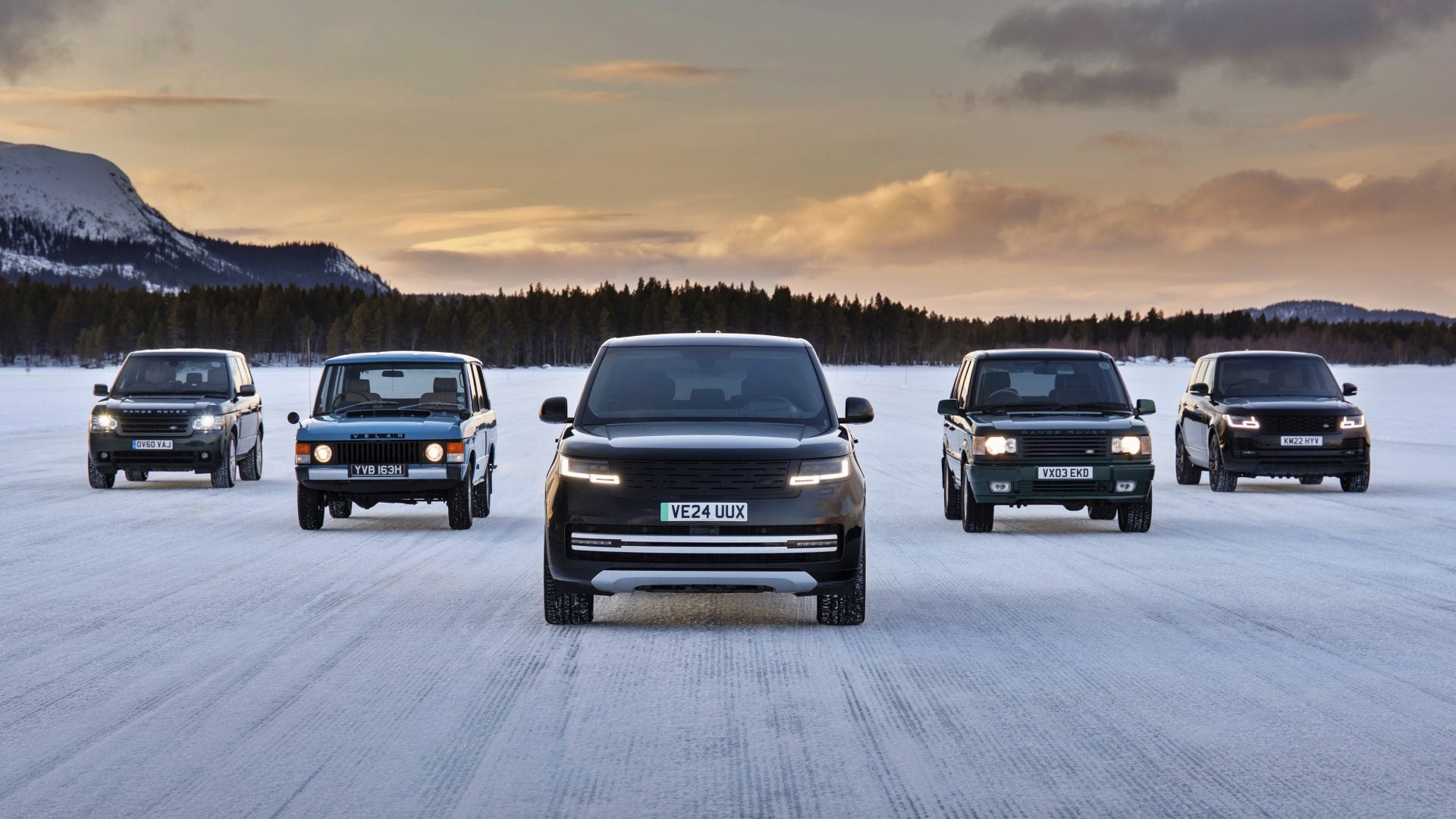 Five Land Rover SUVs of different generations, including the upcoming Range Rover Electric with a 300 mile range, are parked side by side on a snowy landscape at sunset, with mountains and trees in the background.