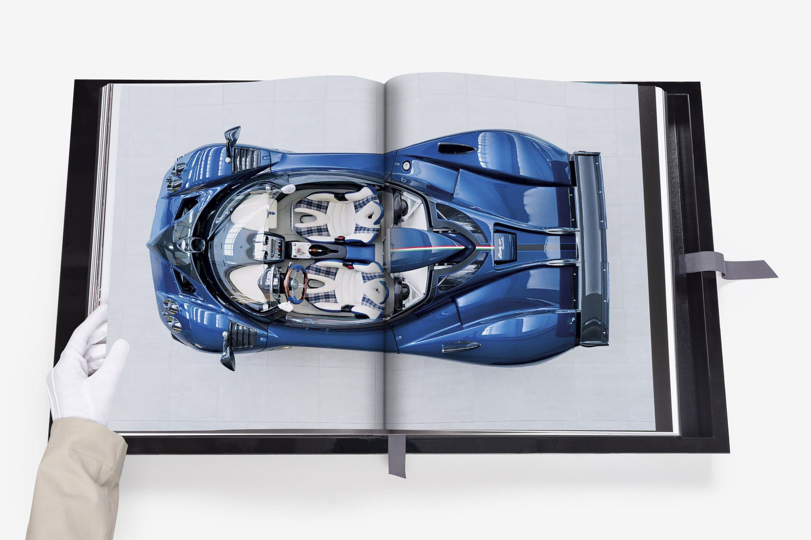 A gloved hand holds open an Assouline book, displaying an image of a blue sports car from a top-down view, showcasing one of its rare cars.