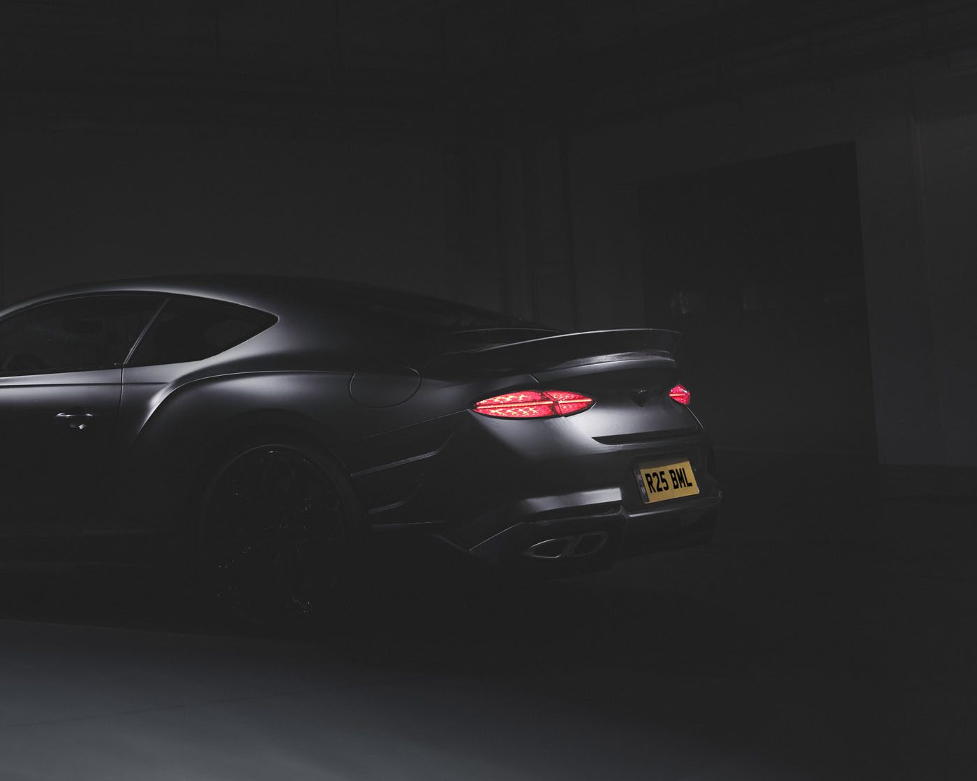 A matte black Bentley sports car is shown from the rear in a dimly lit setting, its taillights glowing and a yellow license plate visible—a rare car from a 100-year-old name.