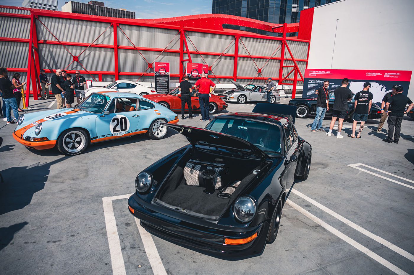 Attendees admire classic Porsche cars, including a black model with its front trunk open, at Luftgekühlt 11—where Bobby Rahal made a Special Guest appearance—against striking red structural elements in the background.