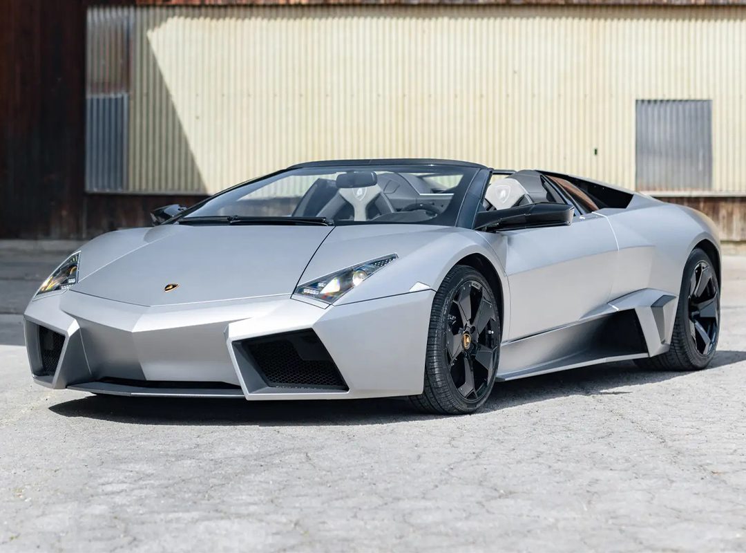 A sleek Lamborghini Revénton Roadster sports car with an angular design is parked on a concrete surface in front of a corrugated metal building, reminiscent of an exclusive find at an RM Sothebys Milan Auction.
