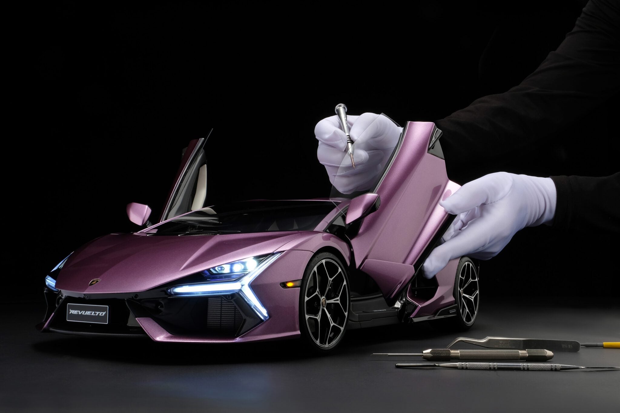 A person in white gloves uses tools to work on a detailed purple Lamborghini Revuelto model with open doors against a black background.