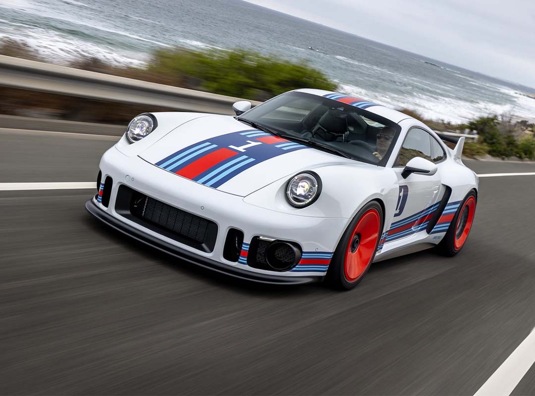 A white Porsche 935 with blue and red racing stripes and red wheels, the ultimate dream car, drives on a scenic coastal road.