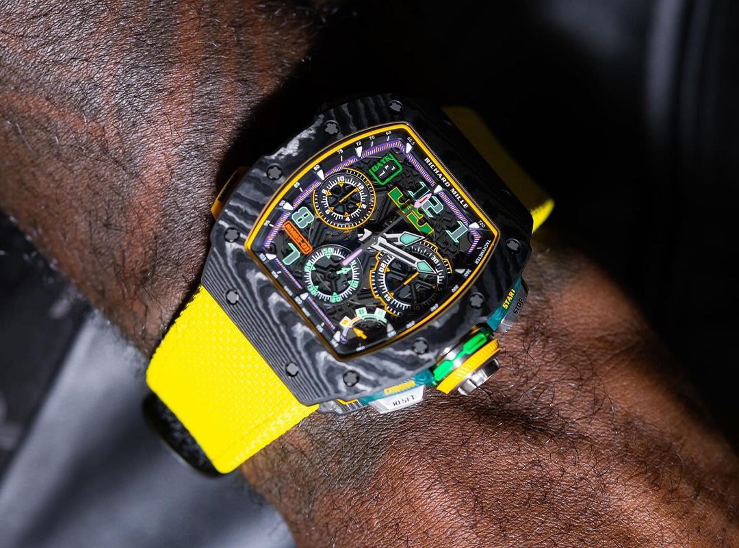 A wrist with a black and yellow high-end sports watch, reminiscent of limited edition watches like the Richard Mille RM65-01, featuring multiple dials and a bright yellow strap.