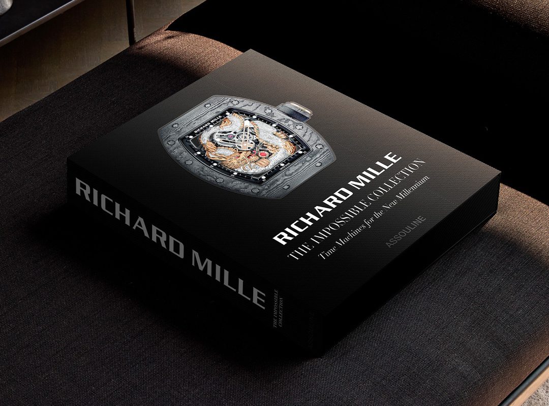 A hardcover coffee table book titled "Richard Mille: The Impossible Collection" by Assouline rests on a dark cushioned surface, featuring a detailed watch on the cover.