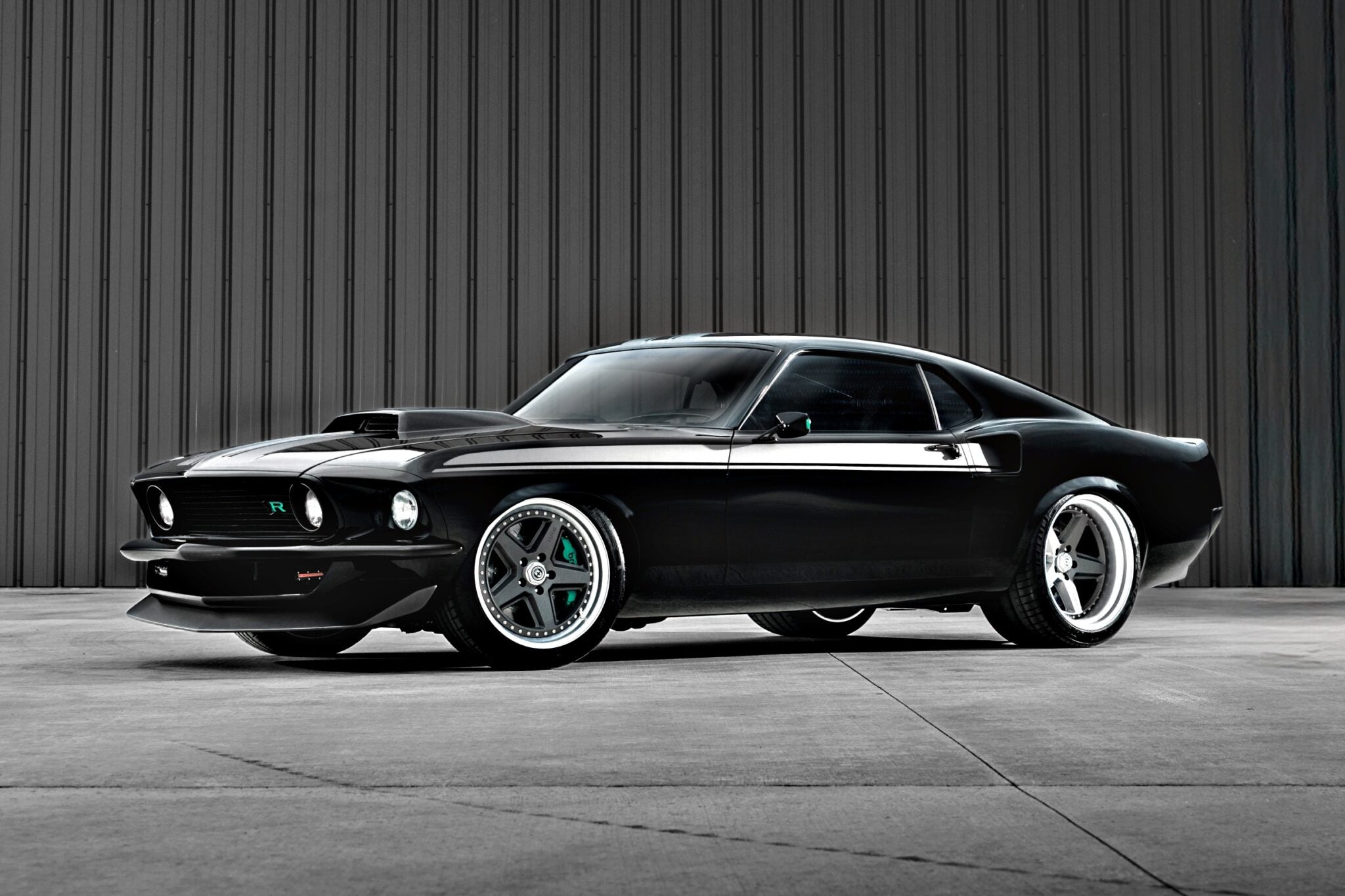 A black classic Ford Mustang with custom modifications, reminiscent of the Ringbrothers' 800-plus horsepower 1969 Mach 1 'KINGPIN' unveiled at the 2025 SEMA, is parked in front of a gray corrugated metal wall.