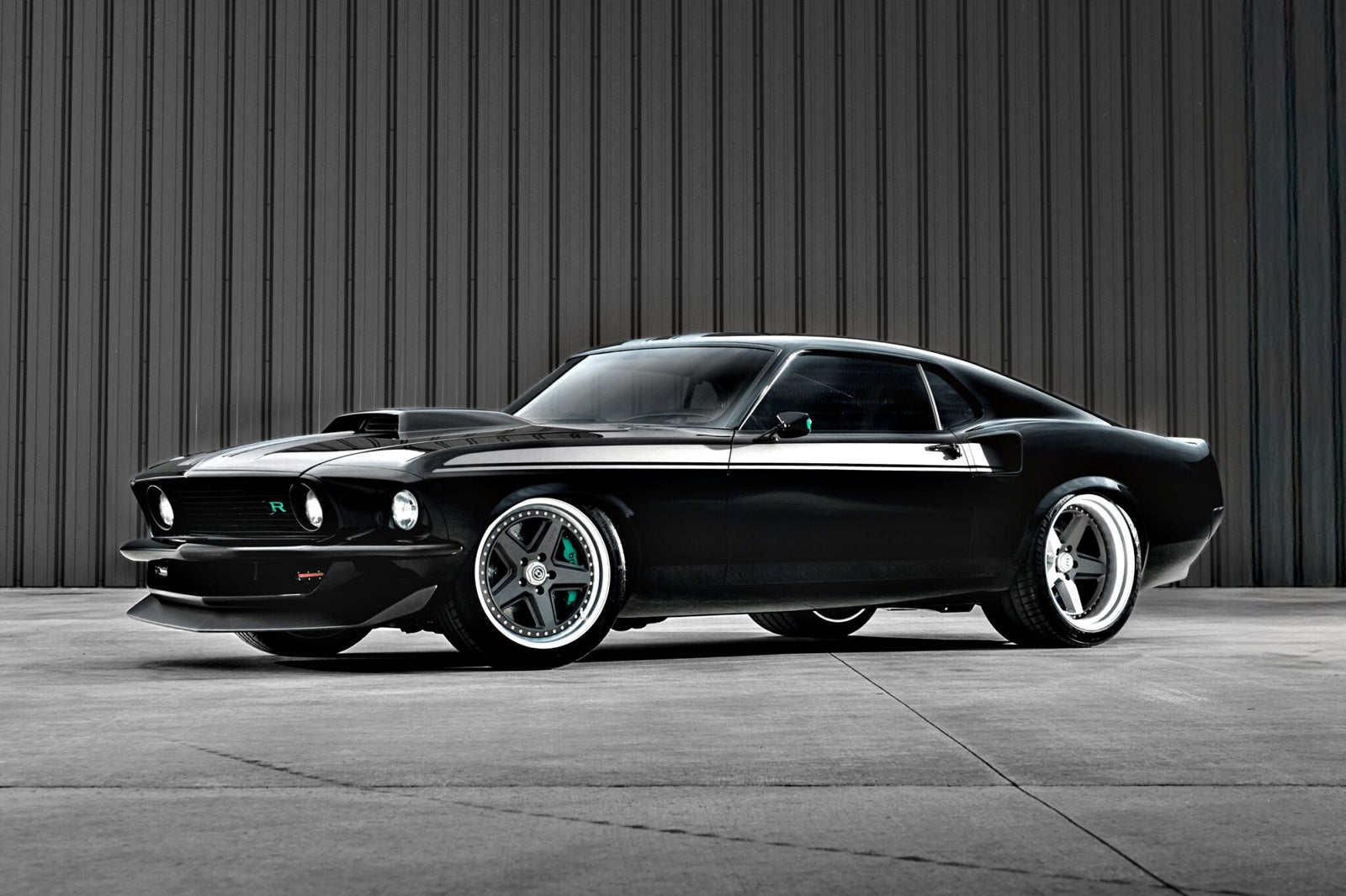 A black classic Ford Mustang with custom modifications, reminiscent of the Ringbrothers' 800-plus horsepower 1969 Mach 1 'KINGPIN' unveiled at the 2025 SEMA, is parked in front of a gray corrugated metal wall.