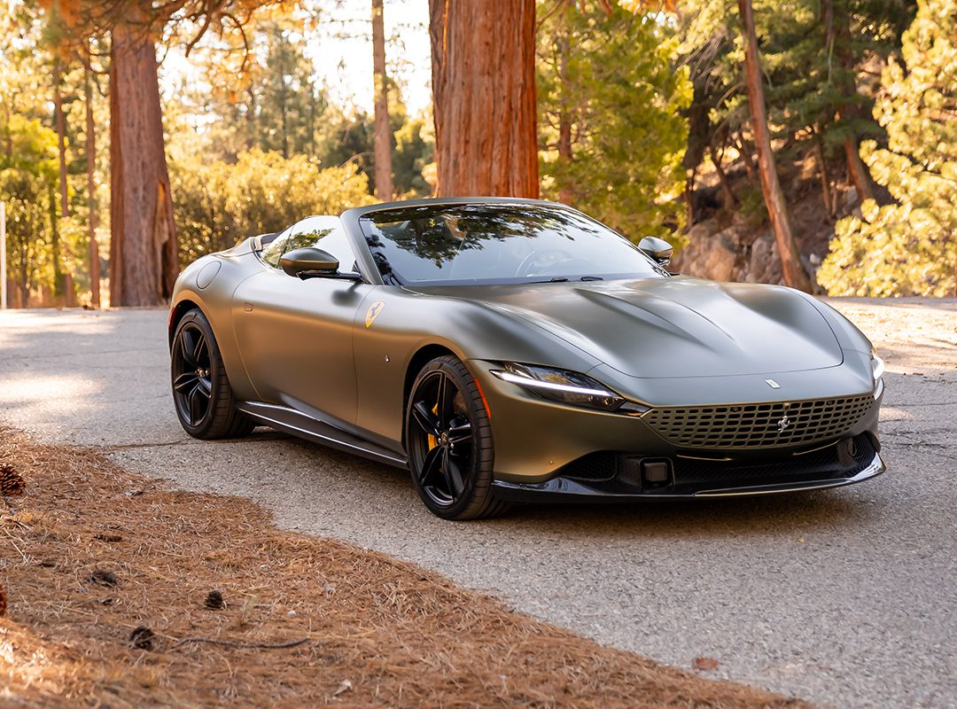 A matte gray Ferrari sports car is gracefully parked on a forest road, with sunlight filtering through the leaves of towering trees, reminiscent of the serene landscapes in California.