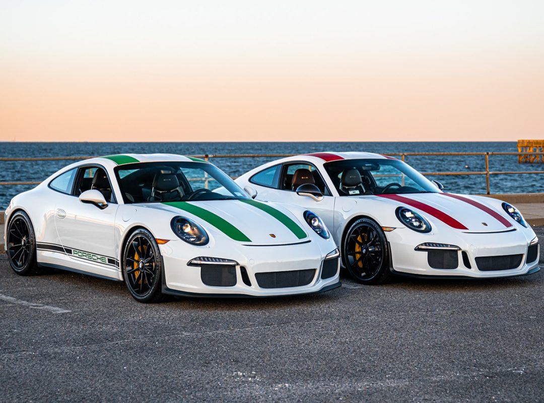 Two white Porsche 911 sports cars—one with green stripes, the other with red—parked side by side by a seaside promenade at sunset, showcase the allure of collector-grade Porsches from Ryan Friedman Motor Cars.