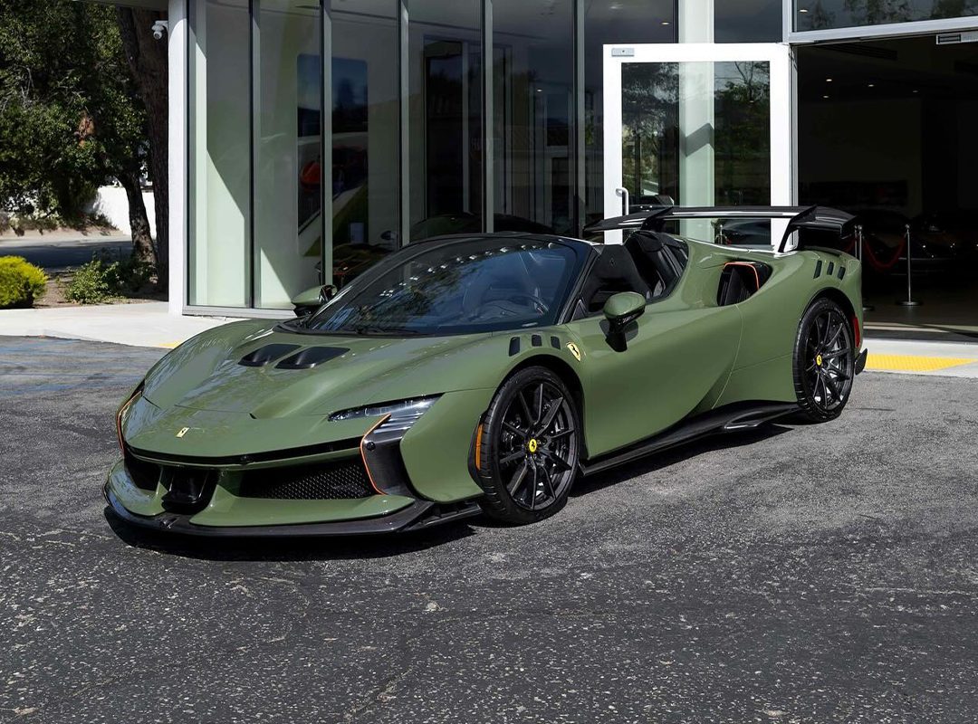 A matte Verde Oliva SF90XX Spider Ferrari convertible is parked outside a modern glass-walled building on a paved surface, capturing the essence of Ferrari Westlake style.