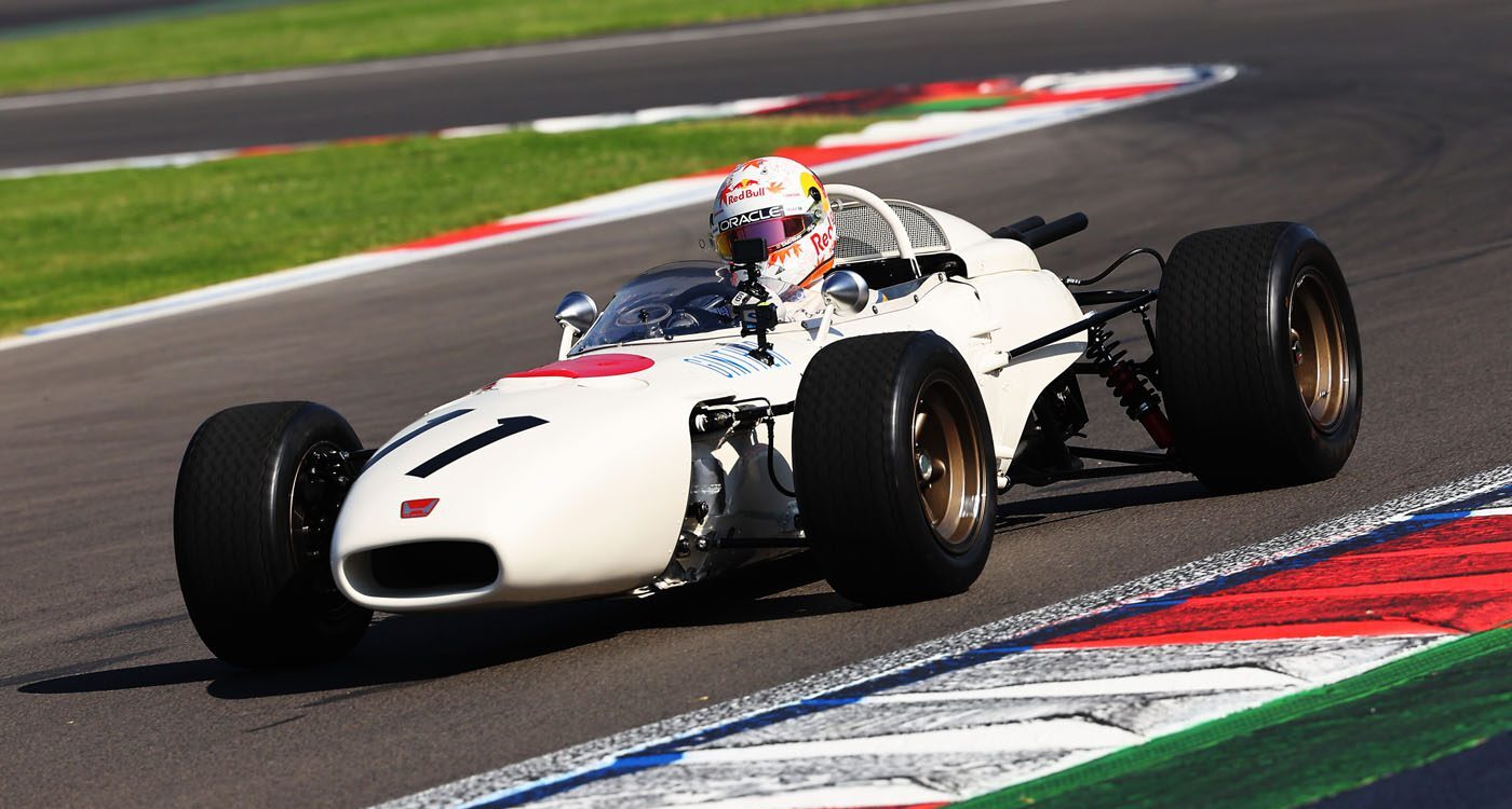 A classic white RA272 open-wheel race car with the number 11, driven by a helmeted driver, takes a turn on a racetrack with colorful curbs, honoring its legacy in motorsport history.