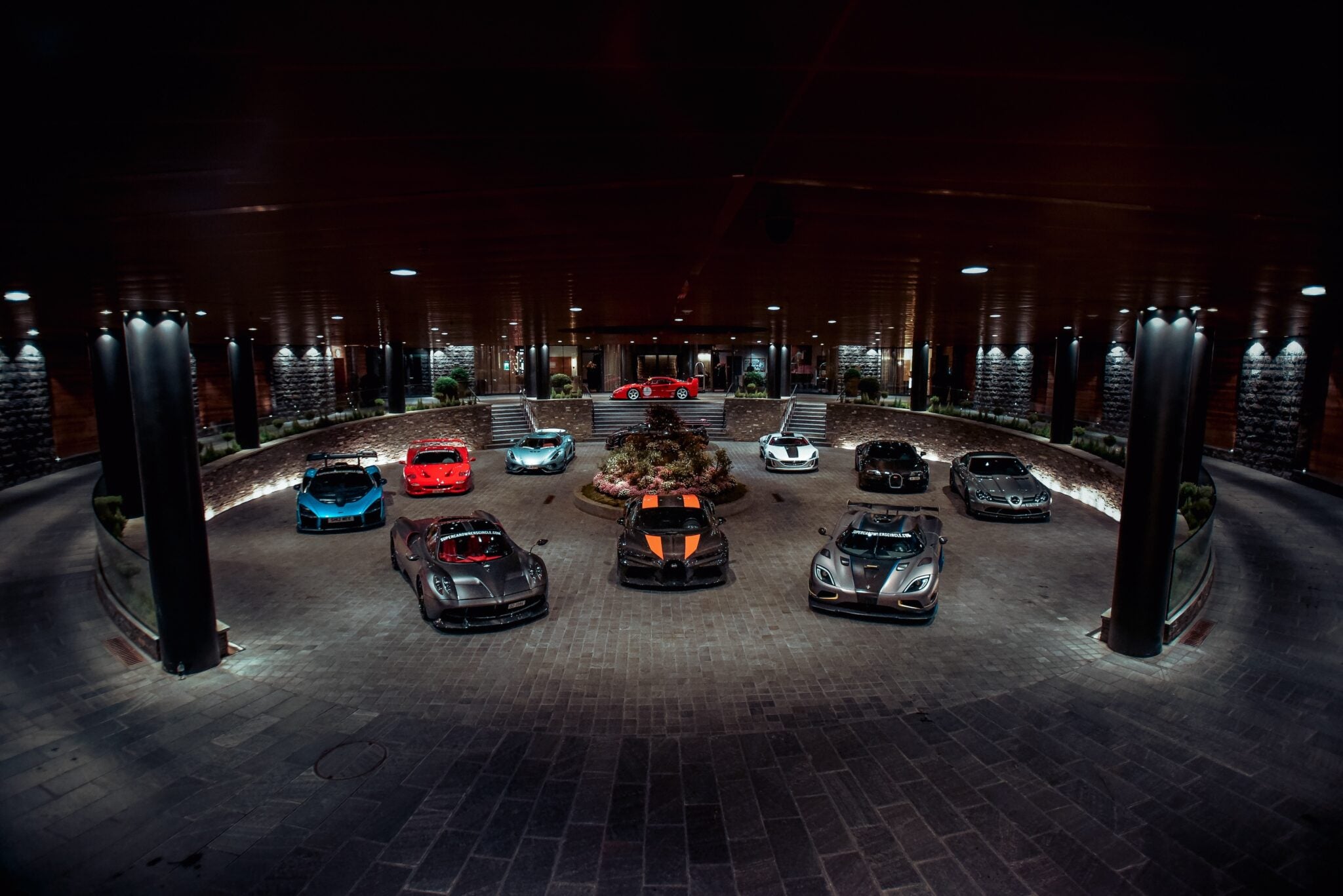 A circular driveway showcases nine parked luxury sports cars, including models in red, blue, silver, and black. The setting is well-lit with columns and spotlights illuminating the scene—a perfect feature for the duPont REGISTRY Supercar Club gathering.