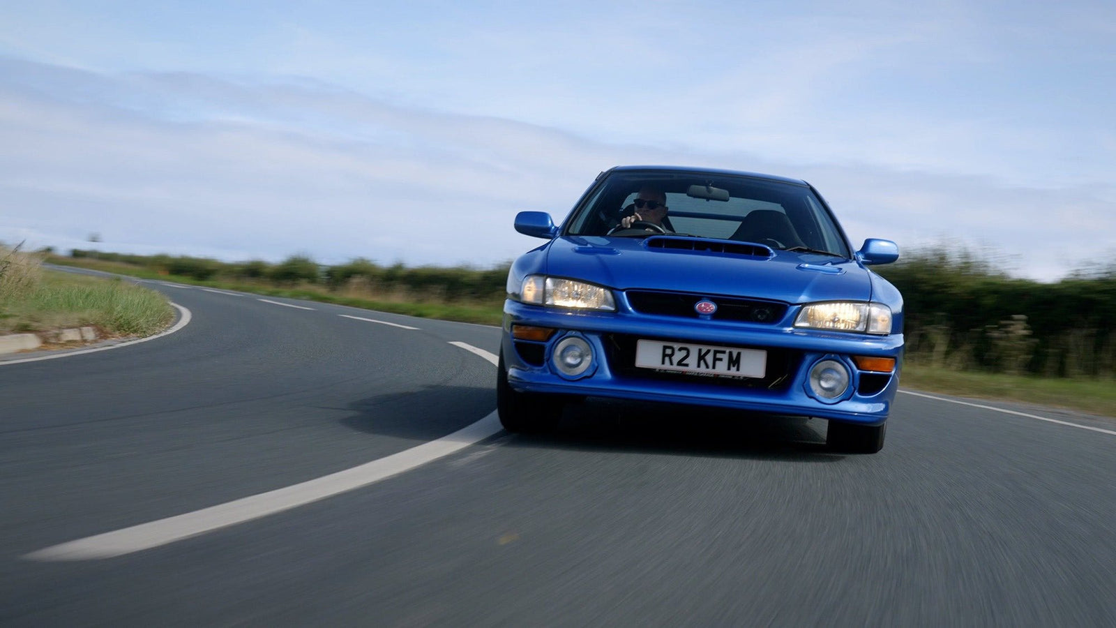 A blue Subaru 22B STi with the license plate "R2 KFW" is driving on a curved, paved road with greenery and a cloudy sky in the background, perfect for a Petrolicious Film Friday feature.