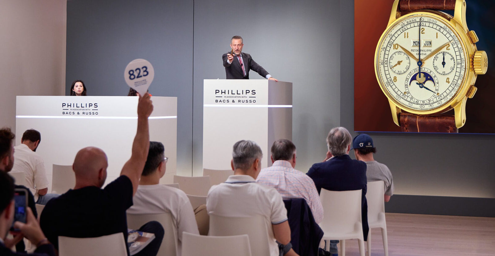 At a New York Watch Auction hosted by Phillips, the auctioneer takes bids on a luxury wristwatch as a participant raises a numbered paddle and others watch the proceedings.