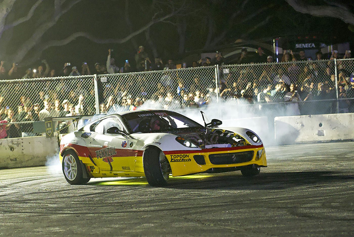 A yellow and white race car drifts around a turn at night during the Monterey Motorsports Festival, producing smoke as a crowd of fans watches from behind a fence.