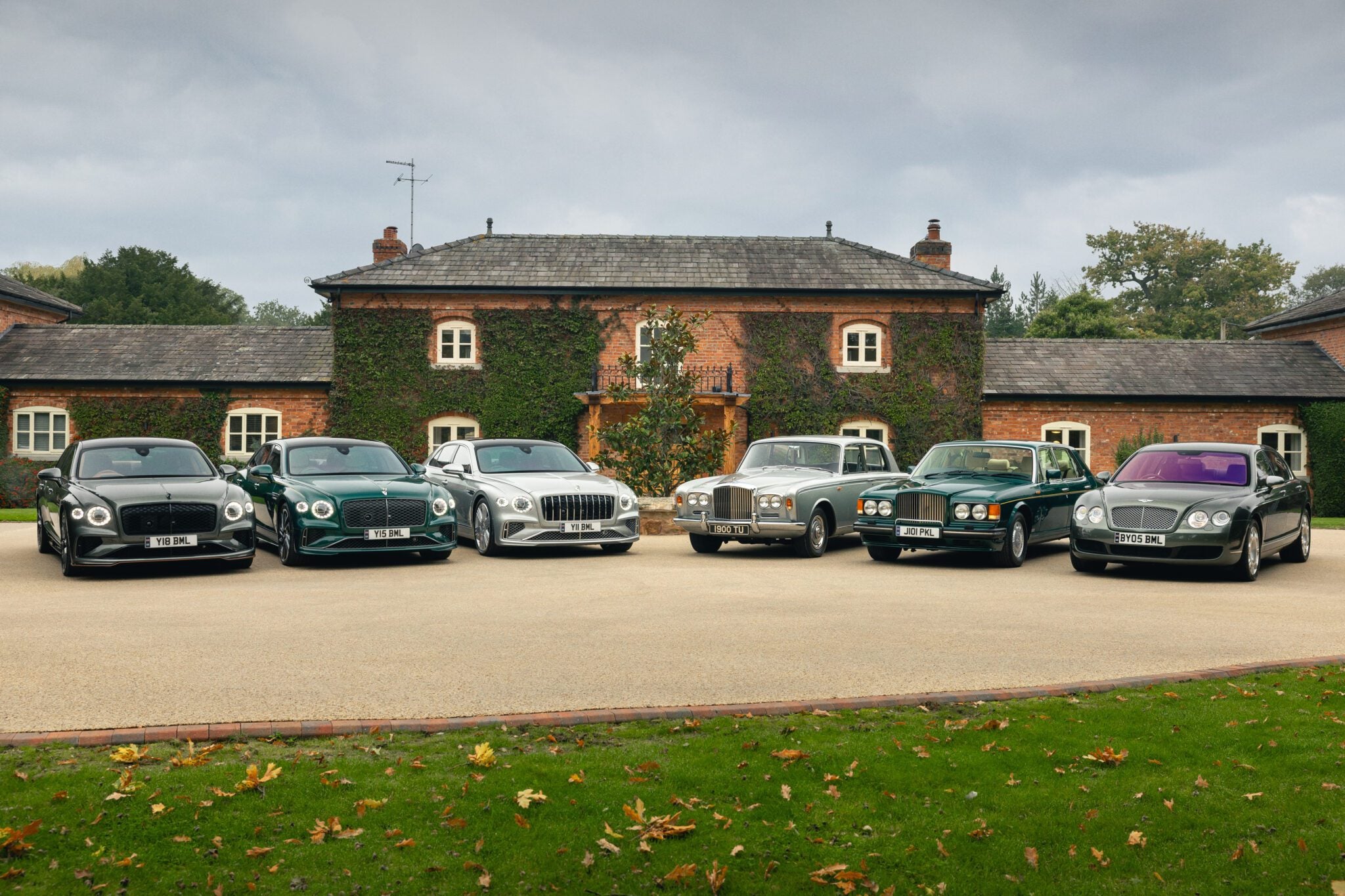 Six legendary sedans are parked in a row on the driveway before a large brick house covered with ivy, including exclusive Bentley Mulliner tributes.