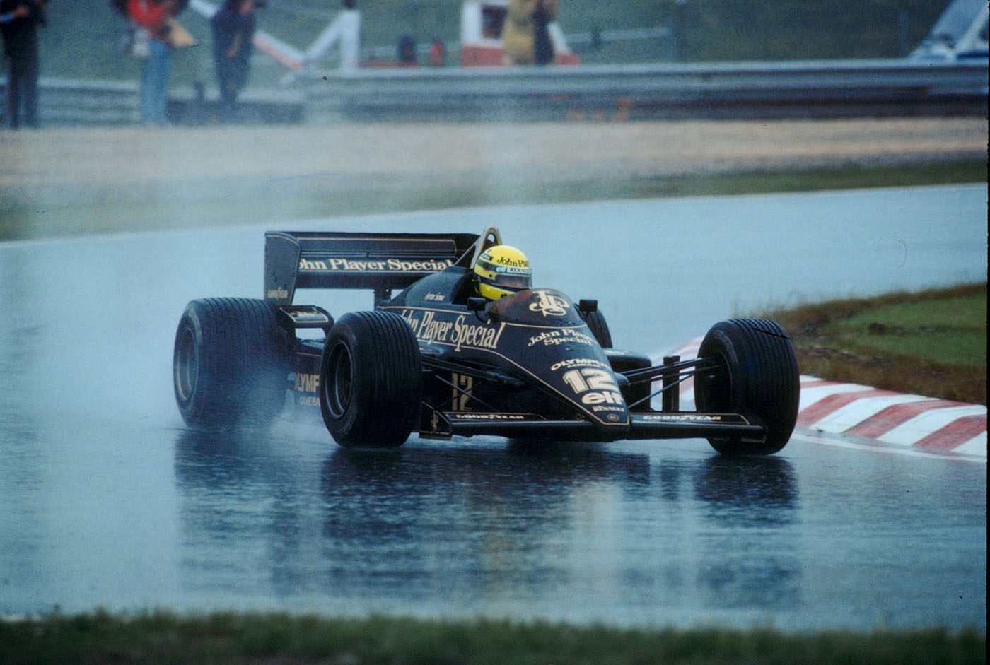 A Formula 1 car drives on a wet racetrack, creating spray behind its tires, with the driver wearing a yellow helmet and the car featuring John Player Special branding.