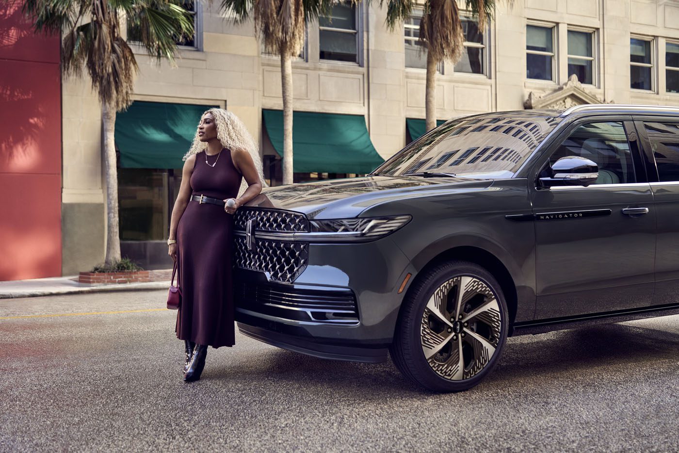 A woman in a brown dress stands beside a gray Lincoln Navigator on a city street. Palm trees sway nearby, and a building with green awnings adds charm to the backdrop.