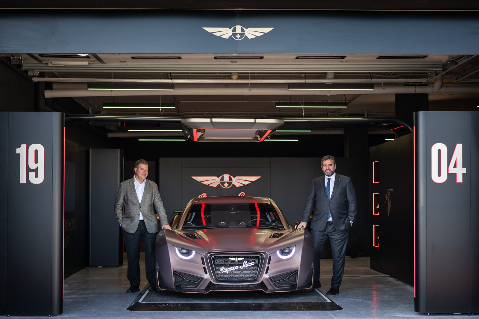 Two men in suits stand beside a sleek, modern Hispano Suiza in a dimly lit garage with the Circuit de Barcelona-Catalunya logo above. The car's front boasts "Hispano Suiza," while the walls are adorned with red and black design elements.