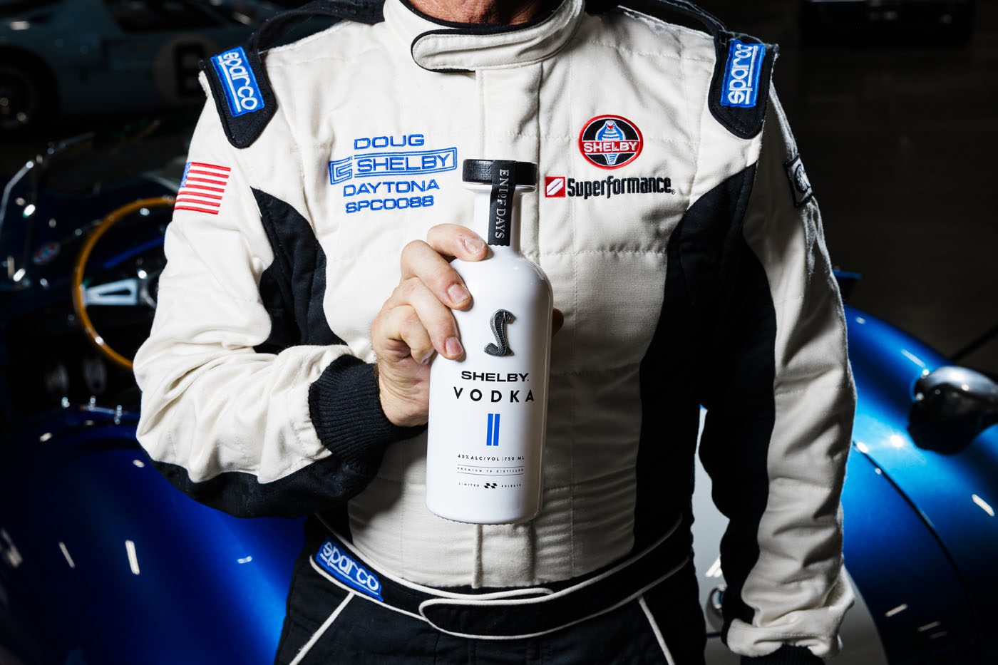 A person in a racing suit holds a bottle of Shelby Vodka, capturing the spirit of a legend in front of a blue sports car.
