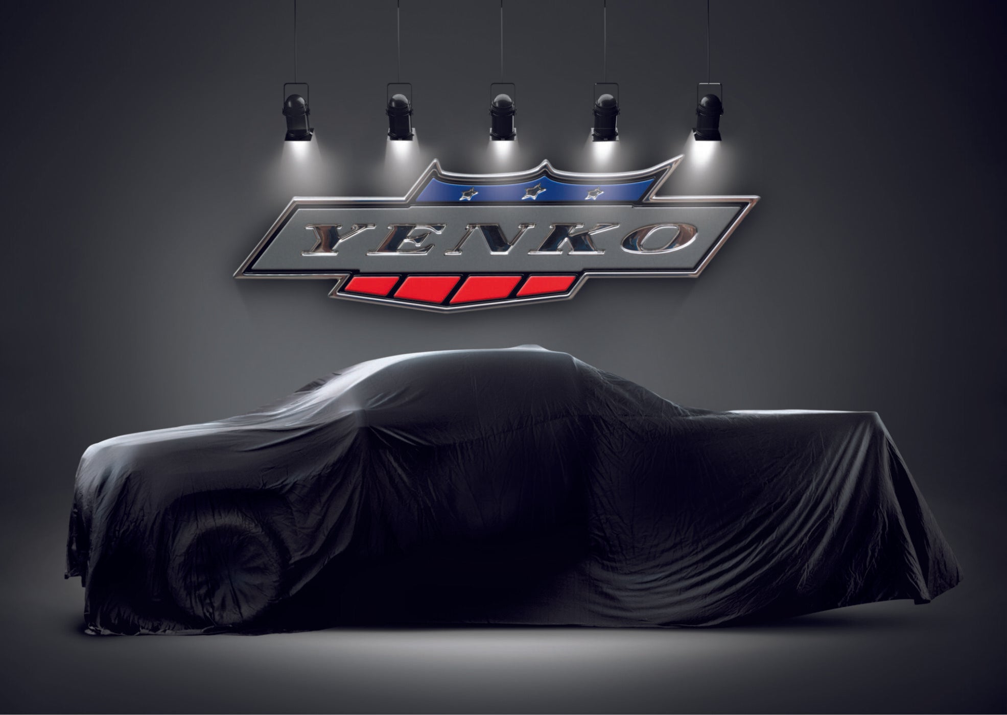 A car covered with a black cloth is displayed under spotlights, with a large Yenko logo mounted on the wall behind it.
