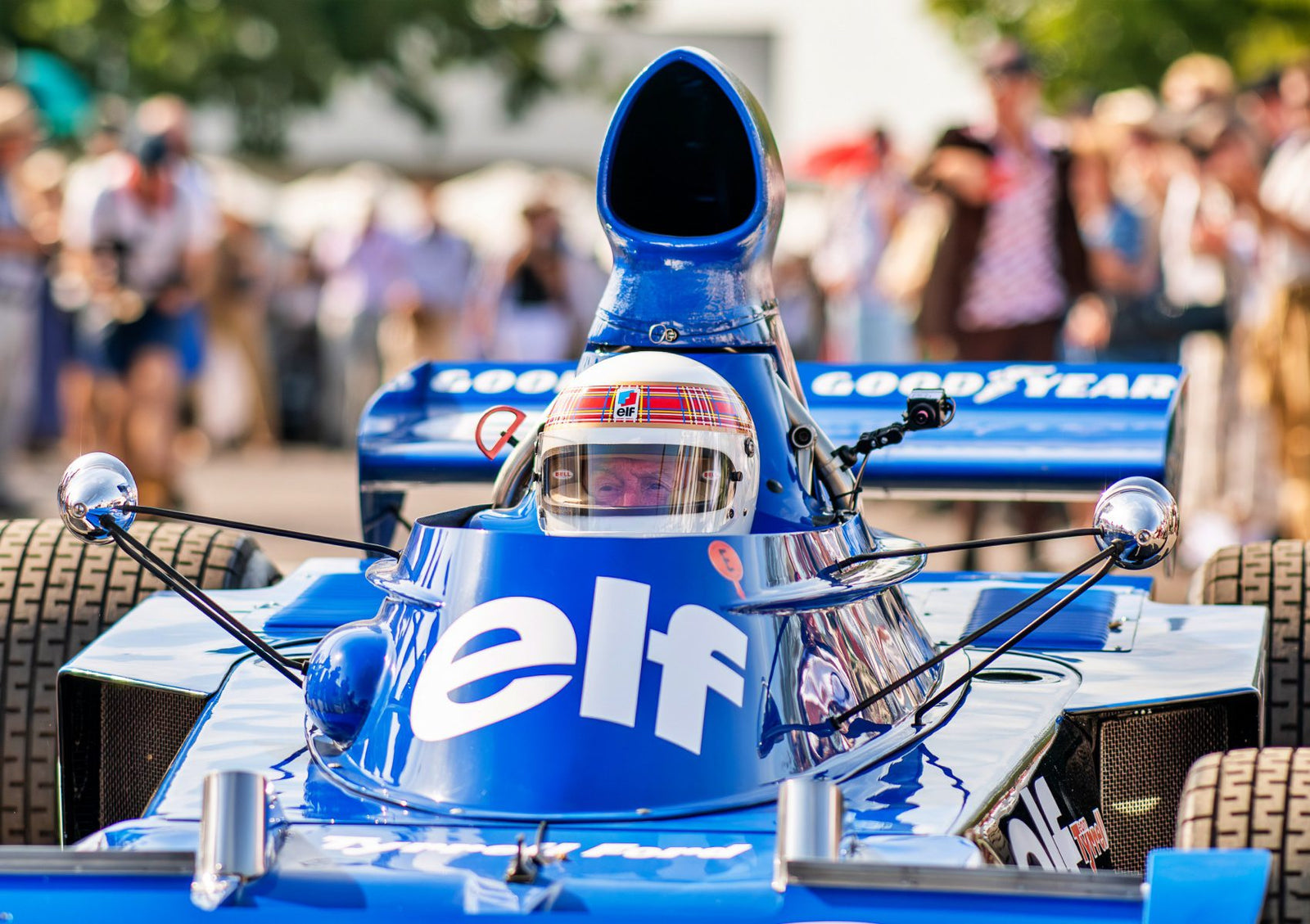 A driver in a blue vintage Formula 1 car with "elf" and "Goodyear" logos prepares to race at the Goodwood Festival, surrounded by a crowd, showcasing one of the era-defining cars of motorsport history.
